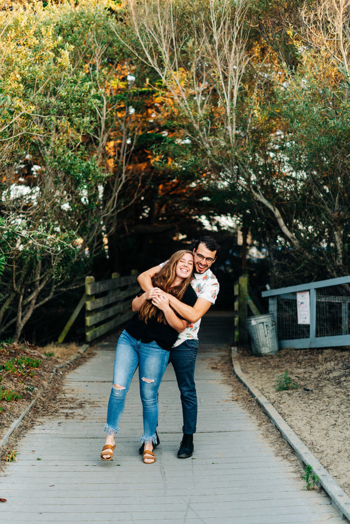 Sunset Engagement Photos at Morro Bay, morro bay wedding photographer; a man hugs his girlfriend from behind, they are in front of some trees on a cement pathway, they are smiling