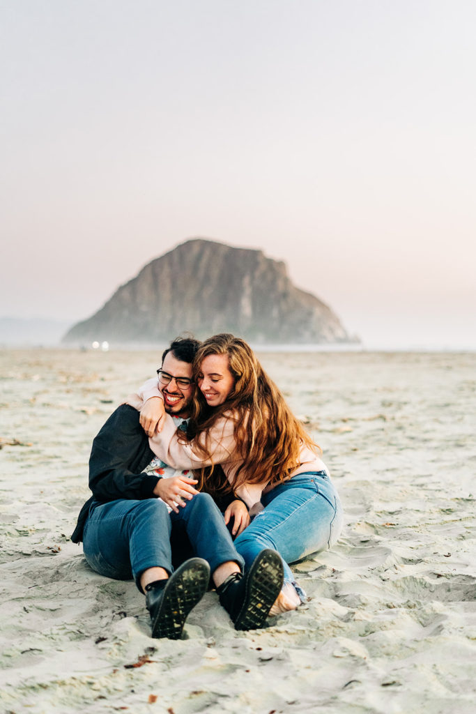 Sunset Engagement Photos at Morro Bay, morro bay wedding photographer; a man and woman couple hug each other as they sit on the beach during sunset in morro bay, california
