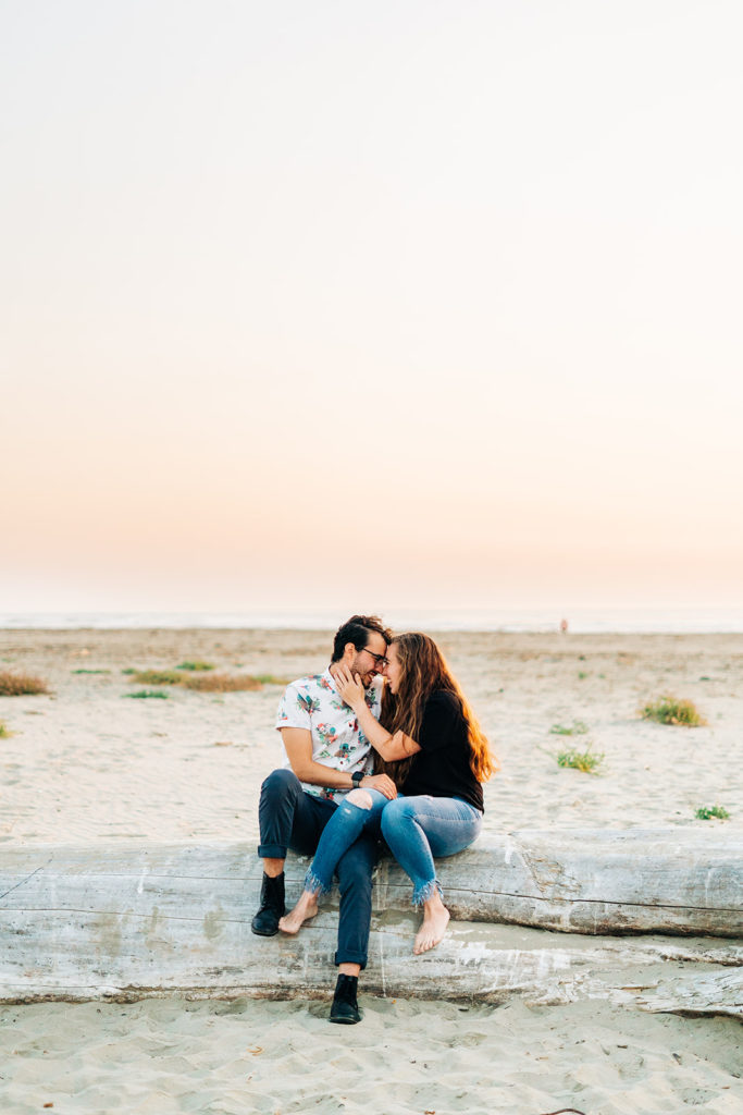 Sunset Engagement Photos at Morro Bay, morro bay wedding photographer; a couple sitting next to each other on a log at the beach, their foreheads are touching. a beautiful, colorful sunset fills the sky.