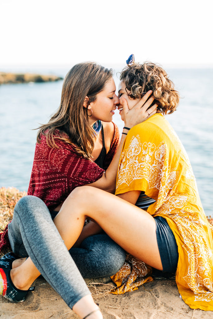 san diego wedding photographer; sunset cliffs engagement photos; two women about to kiss, one is holding the others face. they are sitting on a cliff overlooking the ocean at sunset cliffs in san diego, california