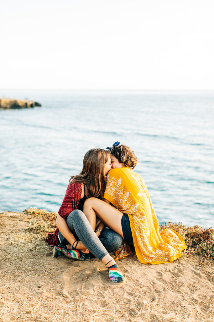 san diego wedding photographer; sunset cliffs engagement photos; two women kissing, they are sitting on the ground on a cliff overlooking the ocean in san diego, ca