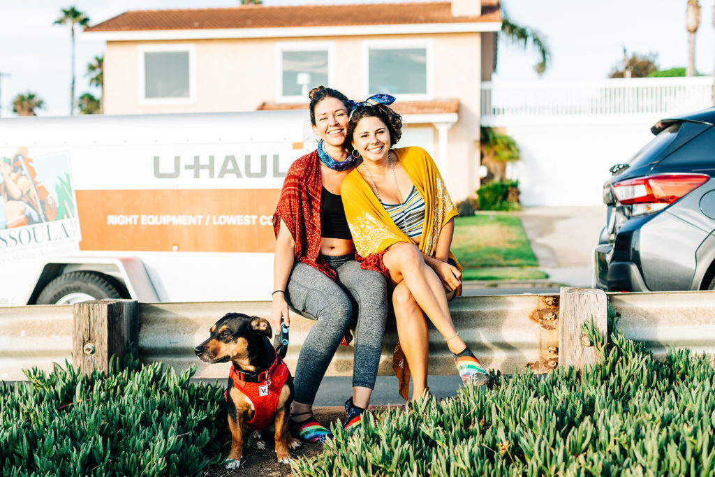 san diego wedding photographer; sunset cliffs engagement photos; two women smiling sitting next to each other in front of a u-haul moving van, they are holding their dog on a leash