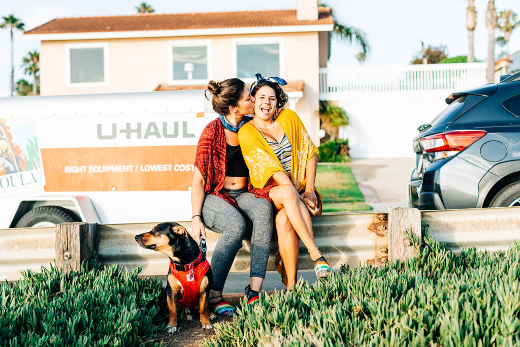san diego wedding photographer; sunset cliffs engagement photos; a woman kissing her girlfriend on the cheek, she is holding her dog on a leash and they are all sitting in front of a u-haul moving trailer