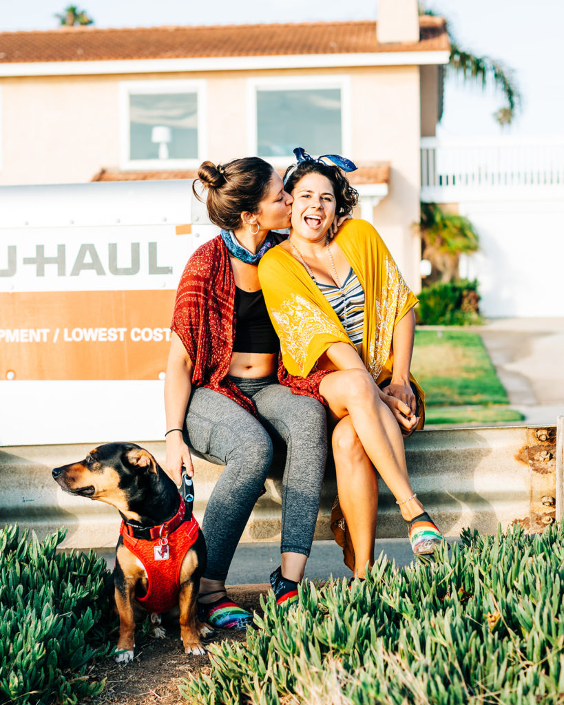 san diego wedding photographer; sunset cliffs engagement photos; a woman kissing her girlfriend on the cheek, they are sitting in front of u-haul moving trailer