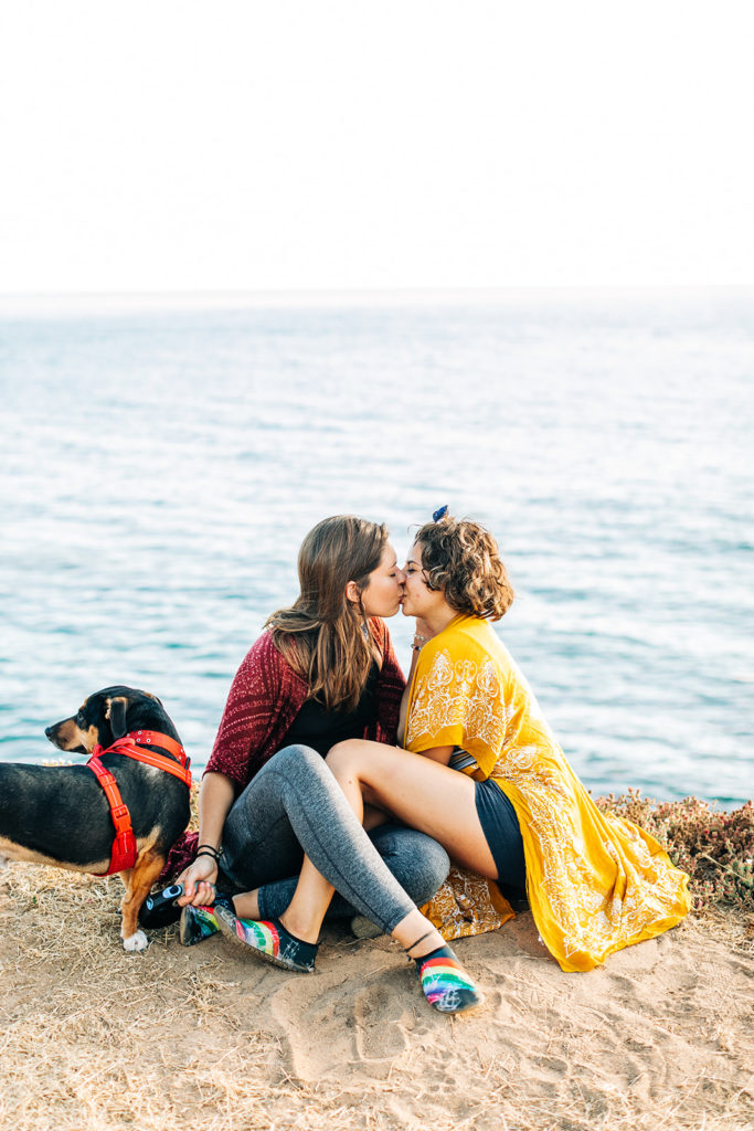 san diego wedding photographer; sunset cliffs engagement photos; two women kissing, they are sitting on the ground on a cliff overlooking the ocean at sunset cliffs in san diego, california. their dog is with them as well.