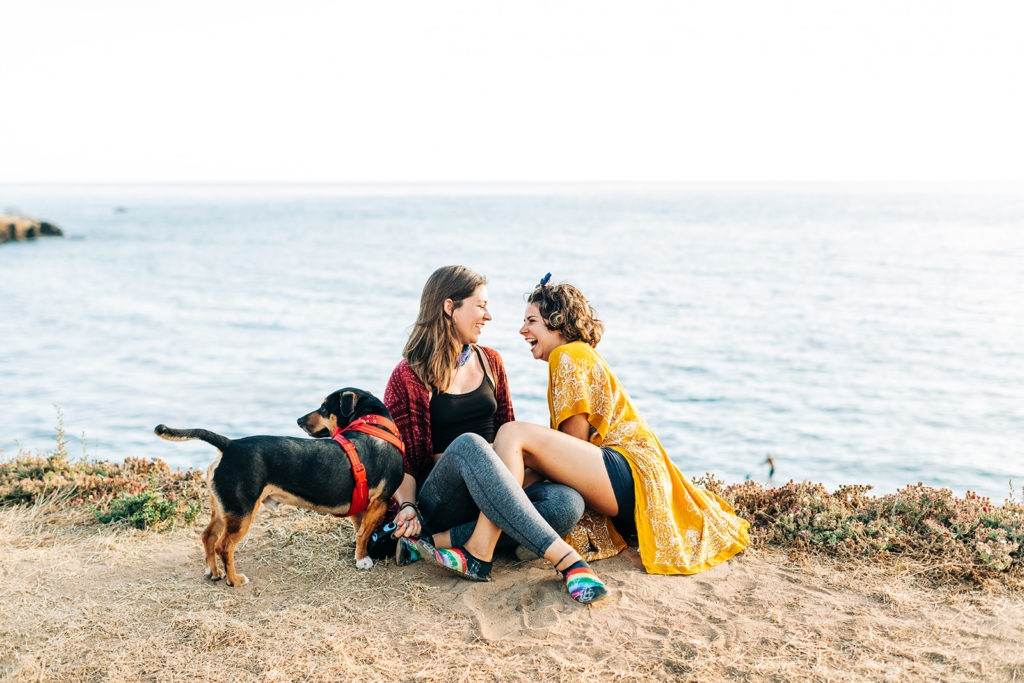 san diego wedding photographer; sunset cliffs engagement photos; two women laughing with each other as they sit close together on a cliff overlooking the ocean at sunset cliffs in san diego, california