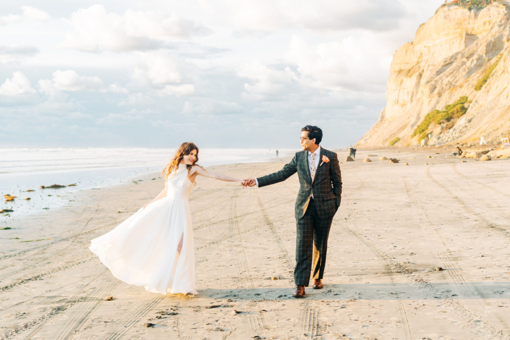 micro wedding in san diego, san diego wedding photographer; a bride and groom hold hands as they walk on the beach at black's beach in san diego