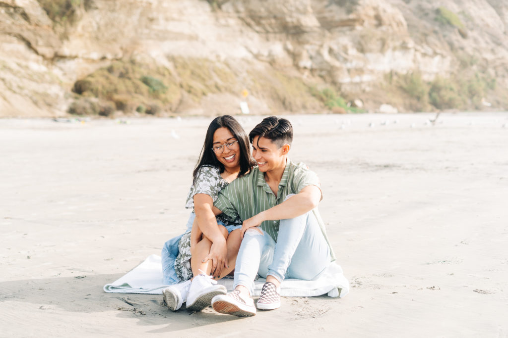 Black's Beach San Diego engagement photos, LGBTQ+ wedding photographer; two women on the beach, they are sitting next to each other on a blanket on the sand