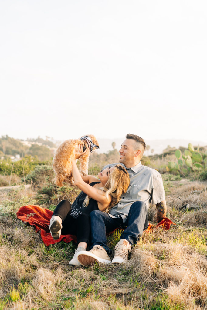 torrey pines gliderport engagement photos, san diego wedding photographer; a man and a woman sitting on a blanket amongst plants while holding up their small brown dog, they are smiling