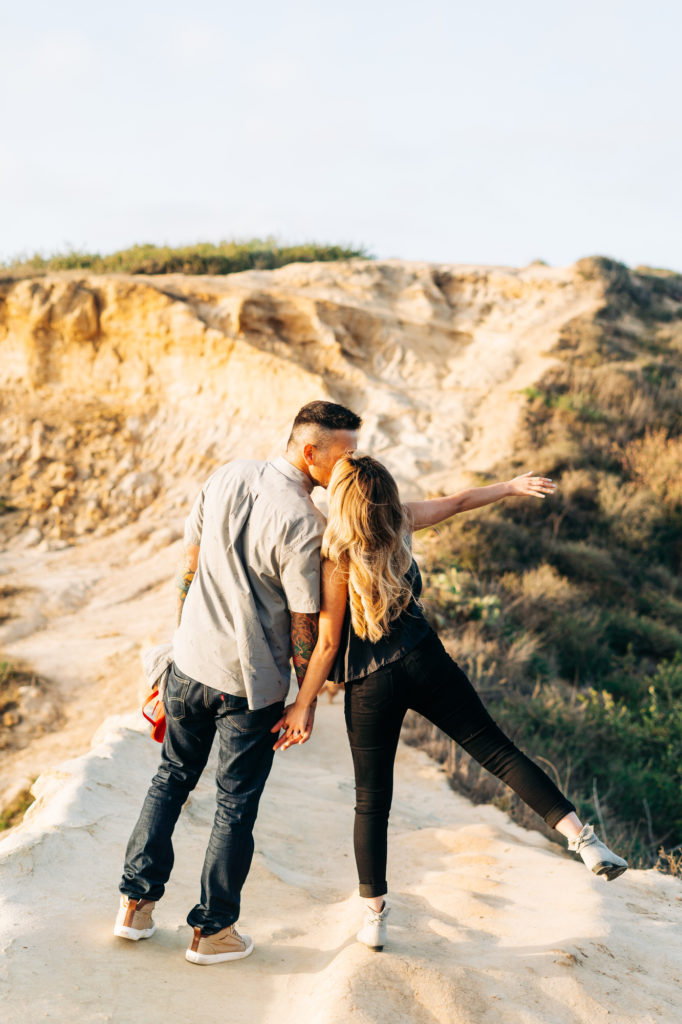 torrey pines gliderport engagement photos, san diego wedding photographer; a man and a woman kiss while walking along a cliff in san diego