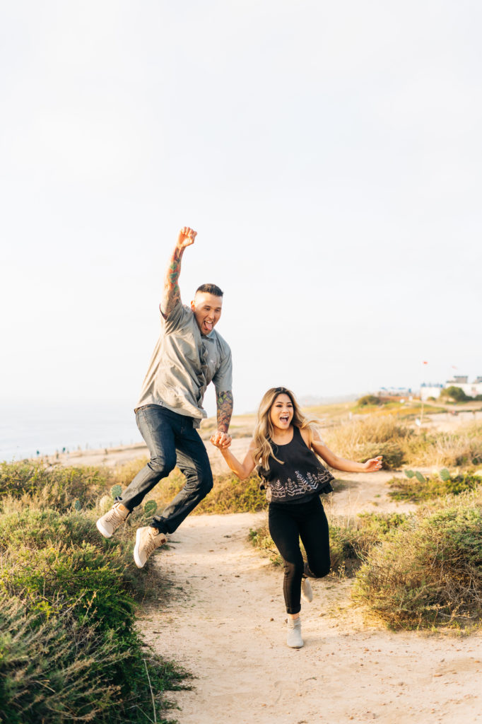 torrey pines gliderport engagement photos, san diego wedding photographer; a man and a woman jump for joy, they are smiling