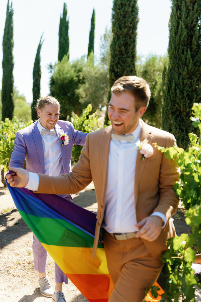 two grooms running with a pride flag on their wedding day; gay wedding in temecula at mount palomar winery