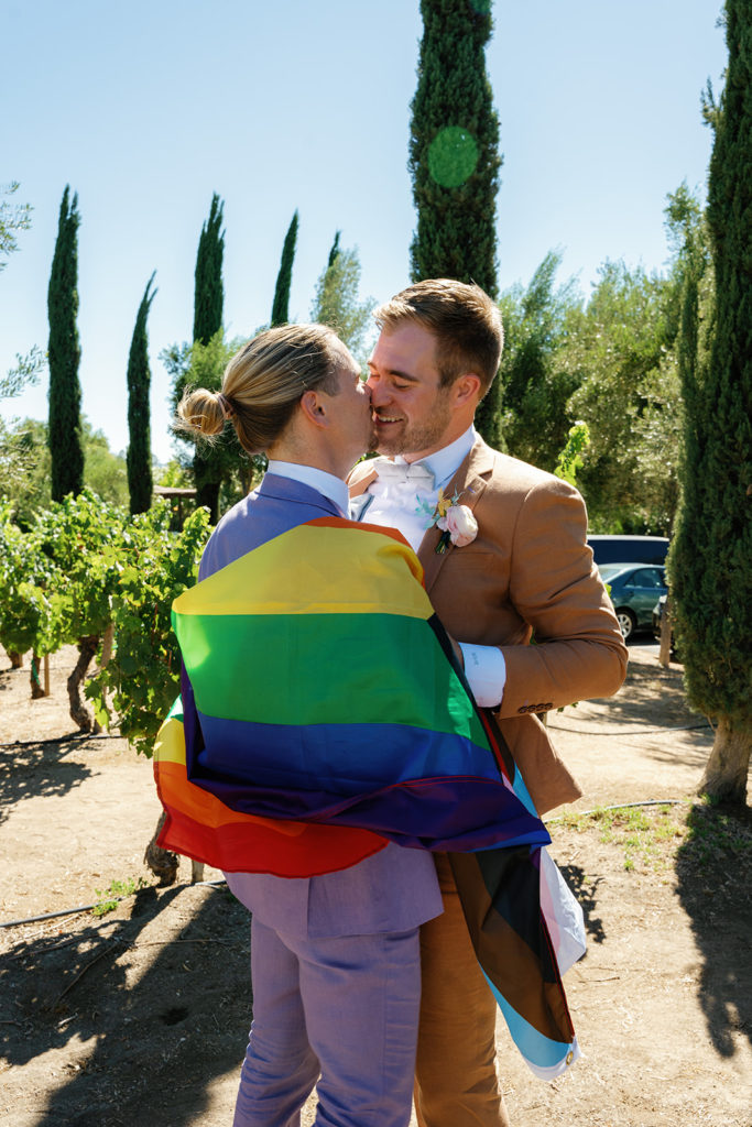 two grooms draped with a pride flag, they are kissing; gay wedding in temecula at mount palomar winery