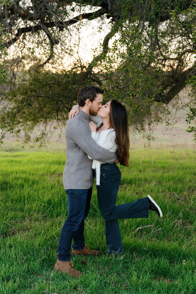 Romantic San Diego Engagement Photos; a man and woman kissing during their engagement photo session. The woman's foot is popped as they kiss.