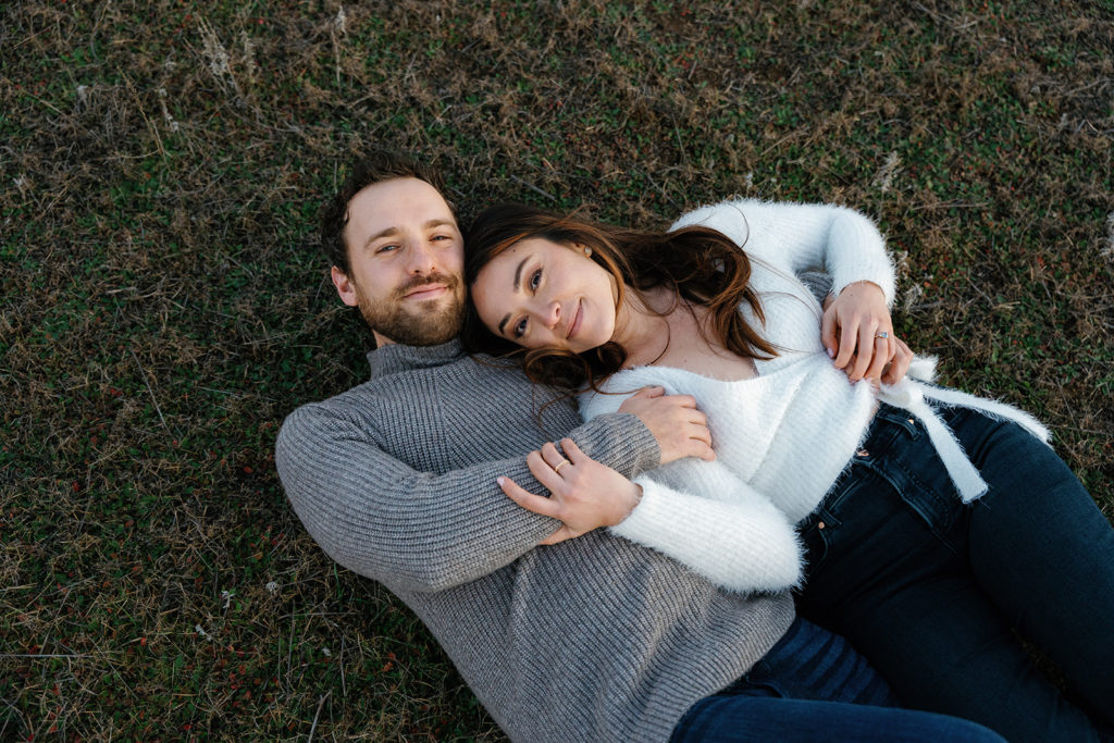 Romantic San Diego Engagement Photos; a man and woman laying down on the grass during their engagement photo session. They are smiling softly at the camera.