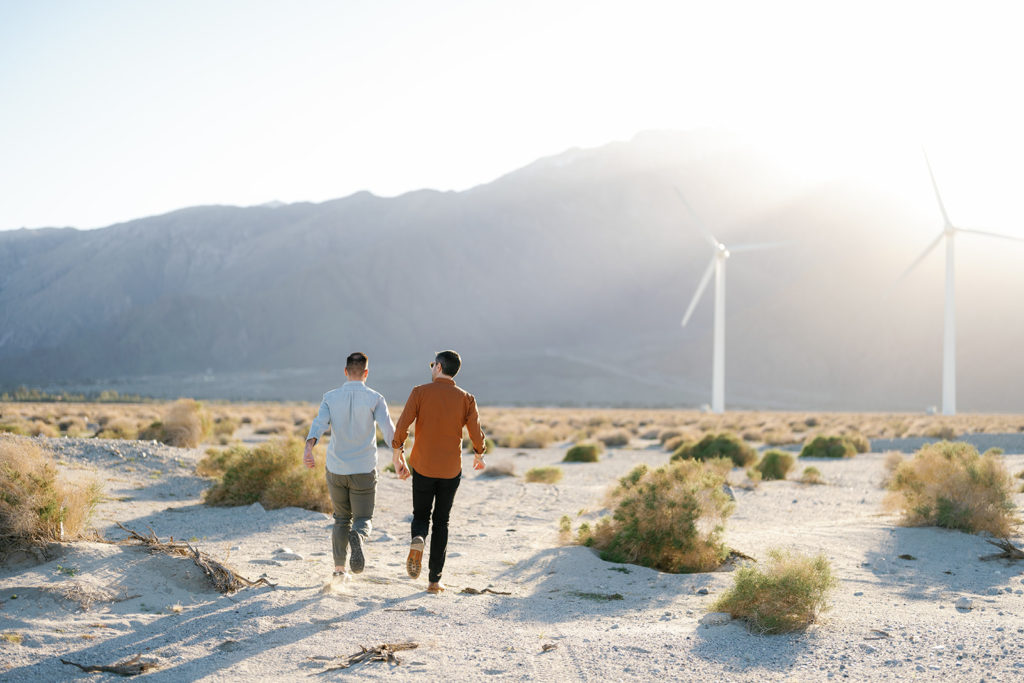 palm springs engagement photos; a gay couple holding hands as they run towards the windmills
