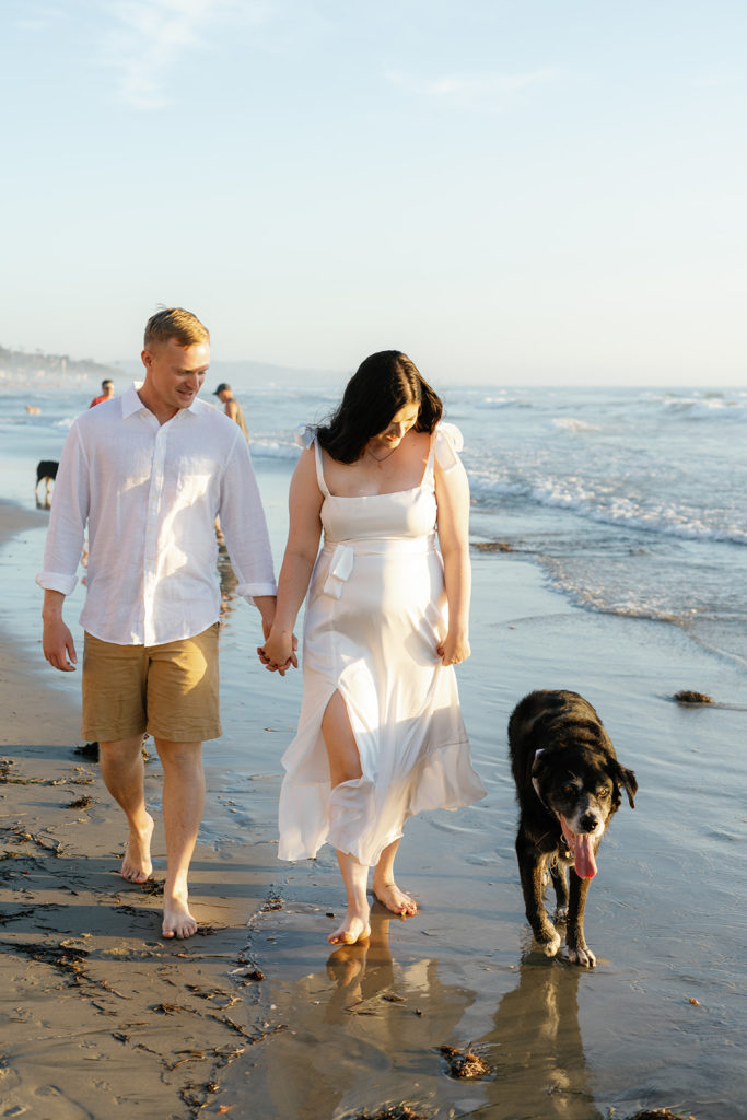 a couple walking on the sandy shore with their dog at their engagement session at del mar dog beach in san diego
