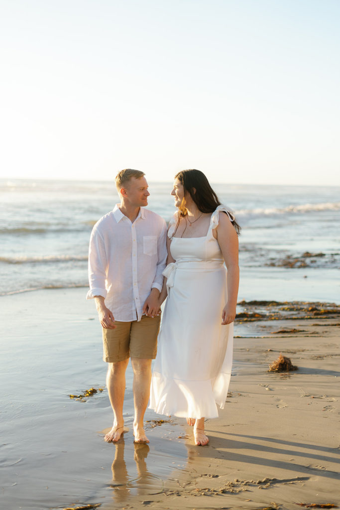 a couple walking on the sandy shore at their engagement session at del mar dog beach in san diego