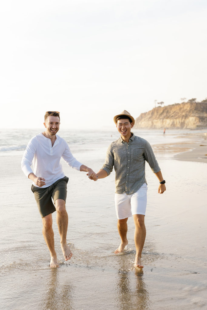 lgbtq+ engagement photos at the beach in san diego; two men holding hands during their engagement session at the beach in san diego, they are running through the water and smiling